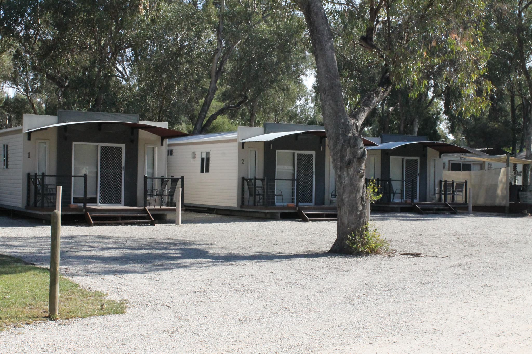 Row of numbered cabins with curved awnings and timber decks under gum trees at Torrumbarry Weir Holiday Park, Patho VIC