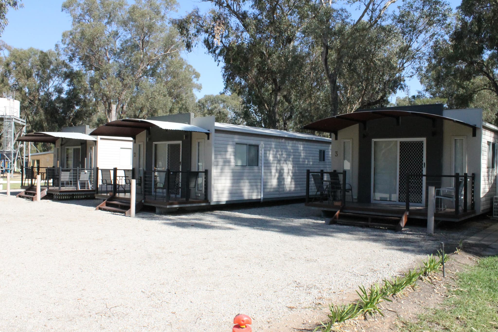 Row of numbered cabins with private decks and curved awnings at Torrumbarry Weir Holiday Park, shaded by gum trees