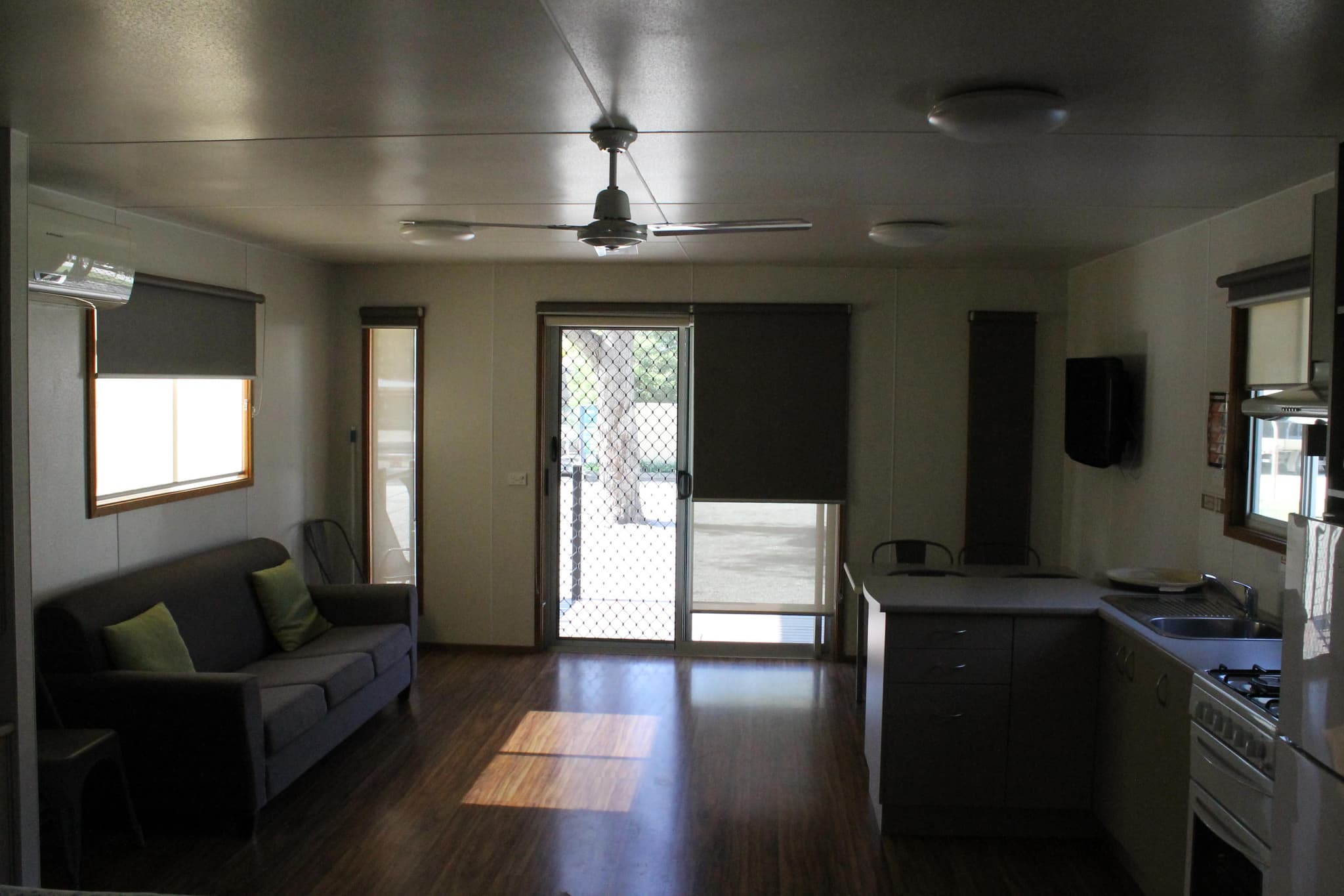 Open plan living area in a cabin at Torrumbarry Weir Holiday Park, with grey sofa, ceiling fan, timber floors and kitchen benchtop