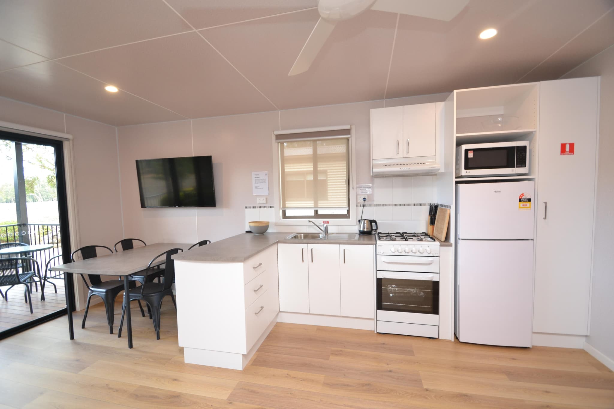 Kitchen and dining area in a Deluxe Riverview Cabin at Torrumbarry Weir Holiday Park, Patho, with white cabinetry and wall mounted TV