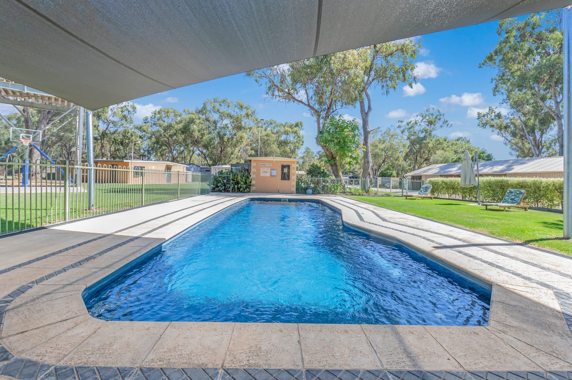 Fenced swimming pool under a large shade sail at Torrumbarry Weir Holiday Park, with lawns, sun loungers and gum trees around it