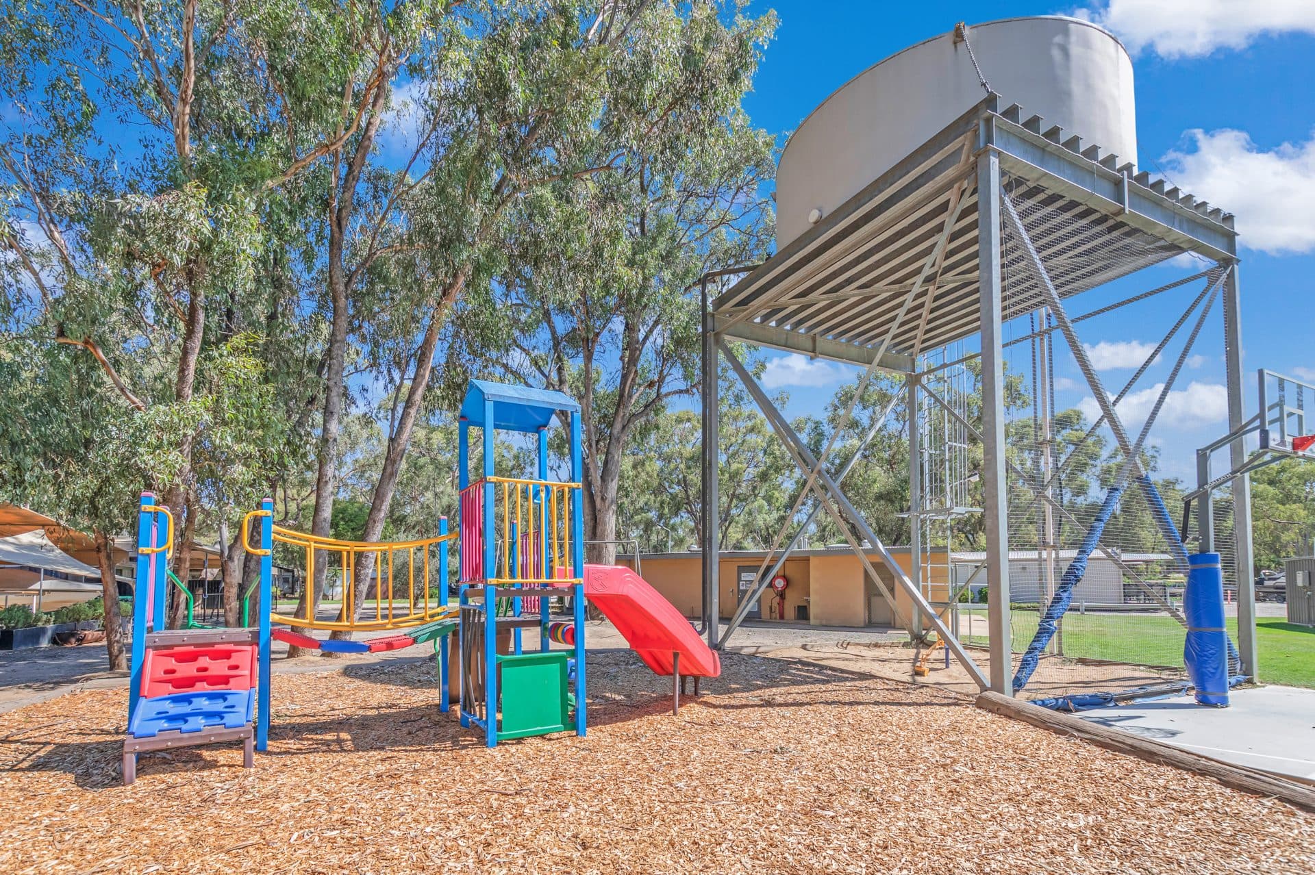 Colourful playground with red slide and bark softfall at Torrumbarry Weir Holiday Park, shaded by gum trees near the water tower