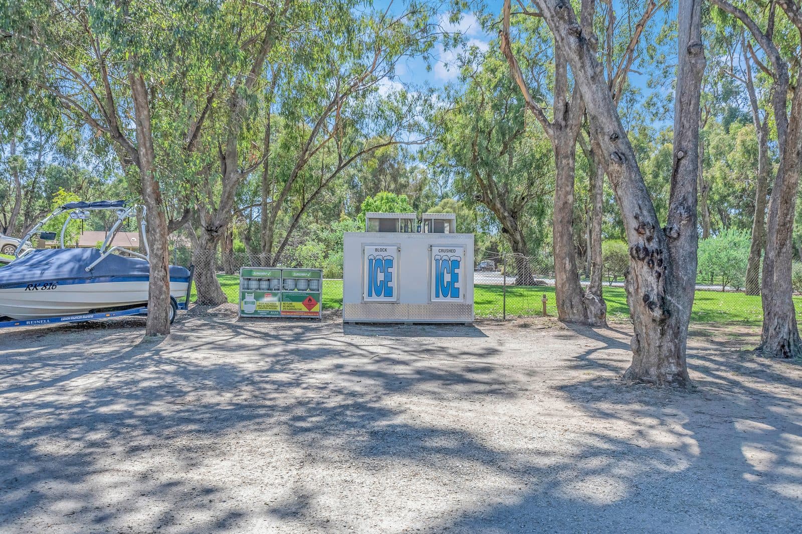 Boat on a trailer parked under red gum trees beside the ice and gas bottle station at Torrumbarry Weir Holiday Park, Patho