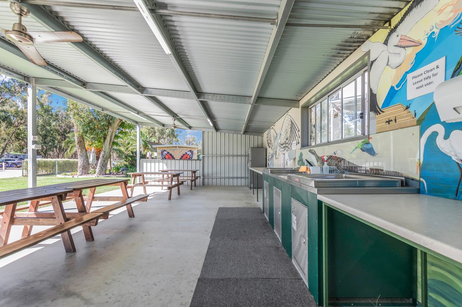 Covered camp kitchen at Torrumbarry Weir Holiday Park with stainless steel benches, sinks and timber picnic tables under a bird mural